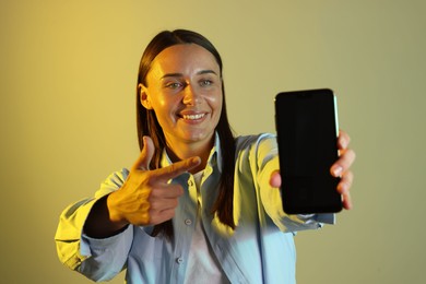 Photo of Smiling woman pointing at smartphone in neon lights against color background