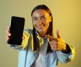 Photo of Smiling woman pointing at smartphone in neon lights against color background