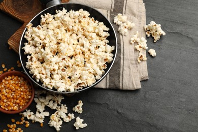 Photo of Tasty popcorn in frying pan and corn kernels on black table, flat lay. Space for text