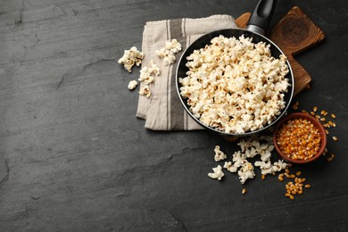 Photo of Tasty popcorn in frying pan and corn kernels on black table, flat lay. Space for text