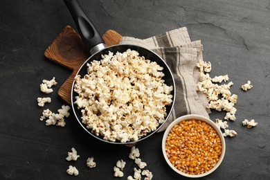 Photo of Tasty popcorn in frying pan and corn kernels on black table, flat lay