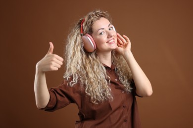 Photo of Happy woman listening to music in headphones and showing thumbs up on brown background