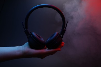 Photo of Woman holding wireless headphones on dark background with color lights and smoke, closeup
