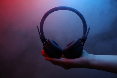 Photo of Woman holding wireless headphones on dark background with color lights and smoke, closeup