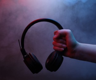 Photo of Woman holding wireless headphones on dark background with color lights and smoke, closeup