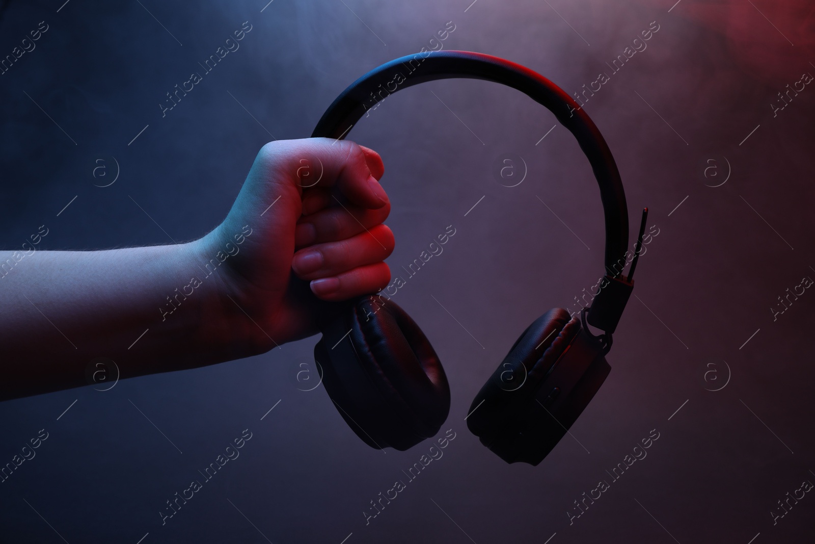 Woman holding wireless headphones on dark background with color lights and smoke, closeup Photo of Woman holding wireless headphones on dark background with color lights and smoke, closeup