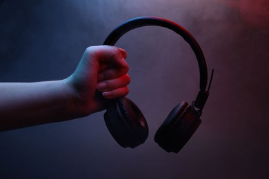 Photo of Woman holding wireless headphones on dark background with color lights and smoke, closeup