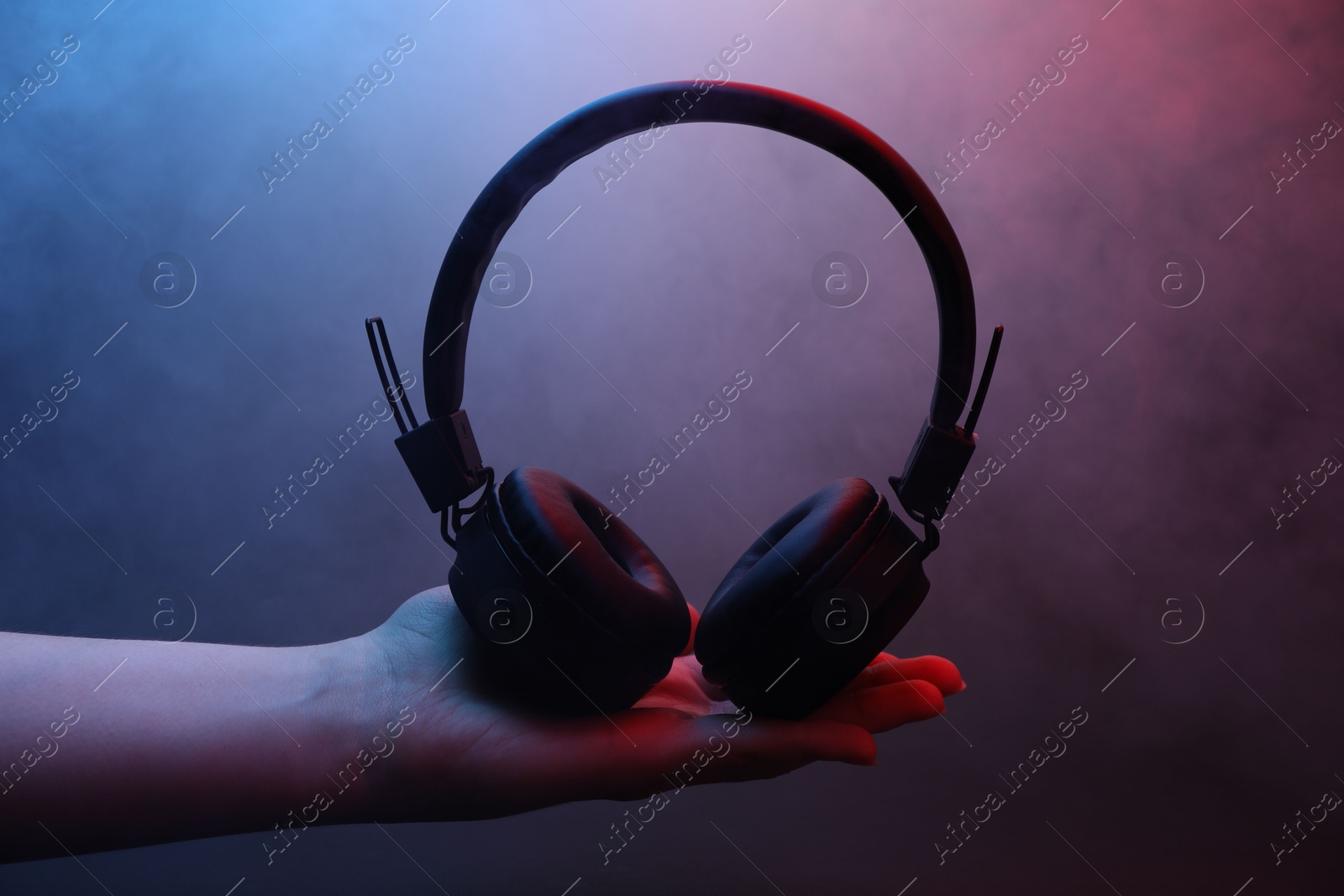 Woman holding wireless headphones on dark background with color lights and smoke, closeup Photo of Woman holding wireless headphones on dark background with color lights and smoke, closeup