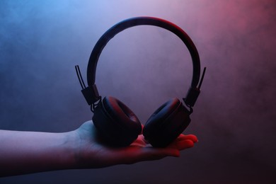 Photo of Woman holding wireless headphones on dark background with color lights and smoke, closeup