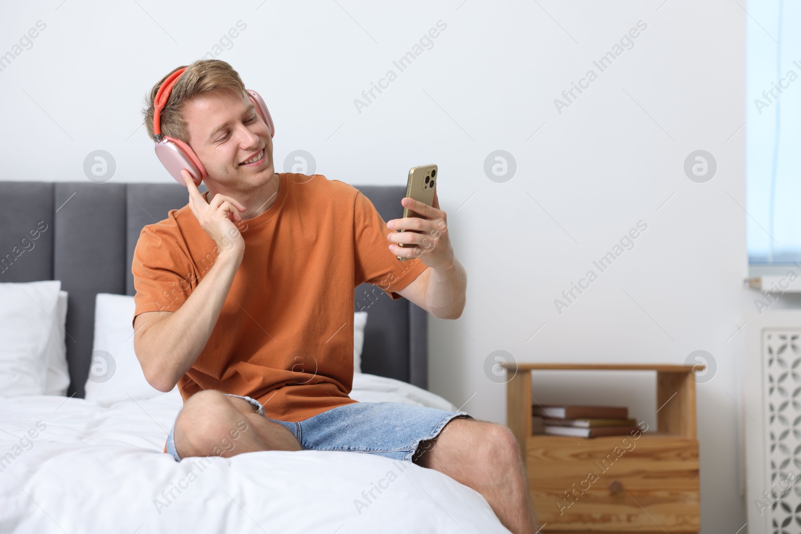 Photo of Happy man with smartphone listening to music in headphones on bed at home. Space for text