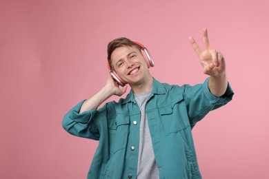 Photo of Happy man listening to music in headphones and showing peace sign on pink background