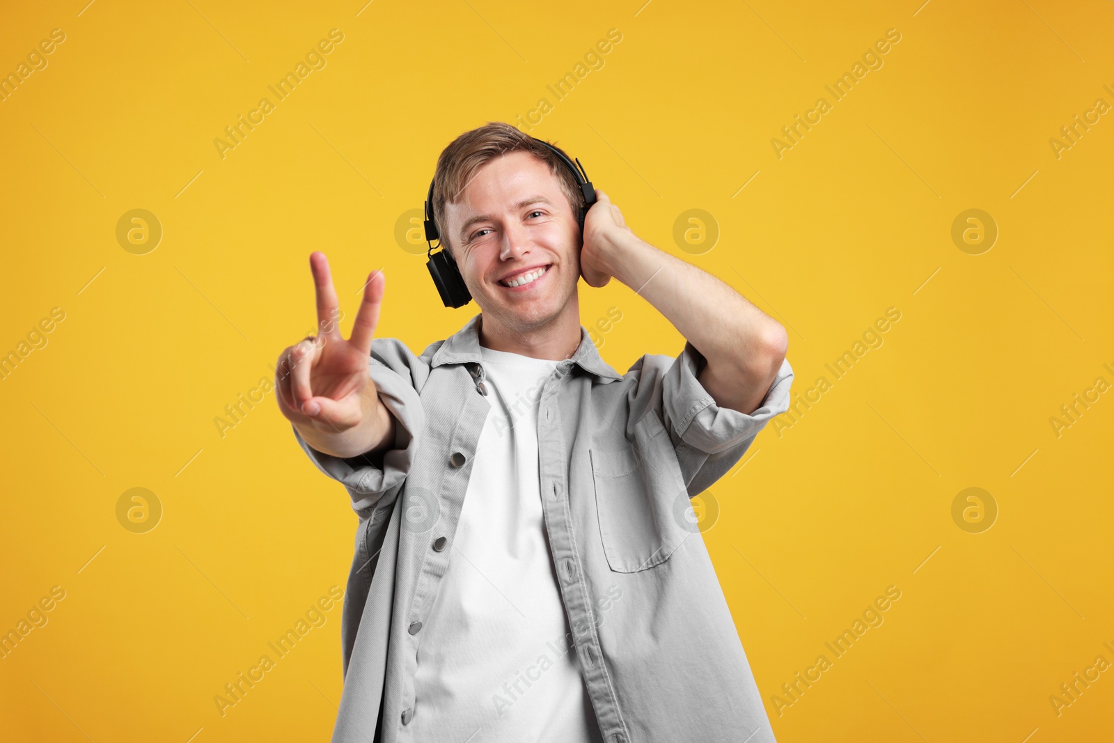 Happy man listening to music in headphones and showing peace sign on yellow background Photo of Happy man listening to music in headphones and showing peace sign on yellow background