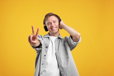 Photo of Happy man listening to music in headphones and showing peace sign on yellow background