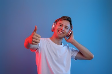 Photo of Happy man listening to music in headphones and showing thumbs up on blue background with neon light
