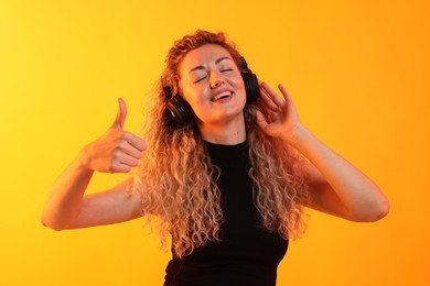 Photo of Happy woman listening to music in headphones and showing thumbs up on yellow background