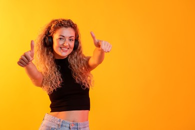 Photo of Happy woman listening to music in headphones and showing thumbs ups on yellow background. Space for text