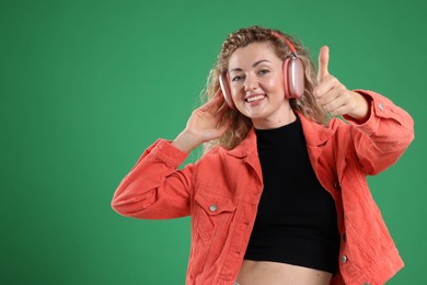 Photo of Happy woman listening to music in headphones and showing thumbs up on green background. Space for text