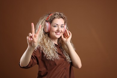 Photo of Happy woman listening to music in headphones and showing peace sign on brown background