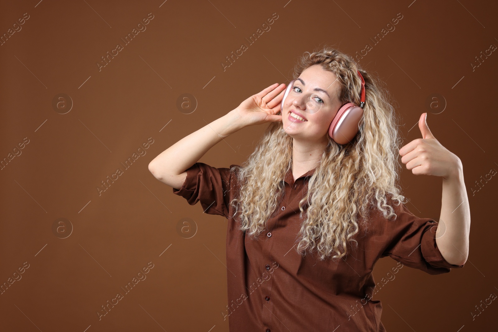 Happy woman listening to music in headphones and showing thumbs up on brown background. Space for text Photo of Happy woman listening to music in headphones and showing thumbs up on brown background. Space for text