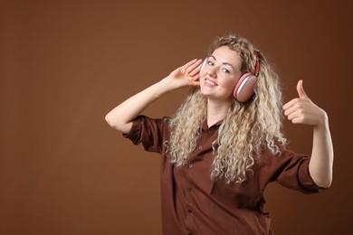 Photo of Happy woman listening to music in headphones and showing thumbs up on brown background. Space for text