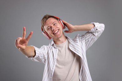 Photo of Happy man listening to music in headphones and showing peace sign on grey background