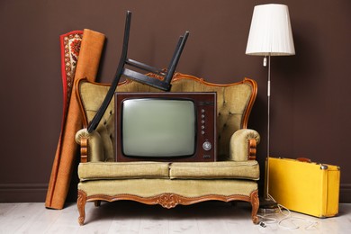 Photo of Retro TV on sofa, chair, suitcase, lamp and carpet near brown wall. Used stuff