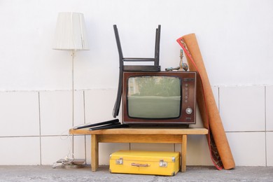 Photo of Retro TV on table, chair, lamp, carpet and suitcase near white wall outdoors. Used stuff
