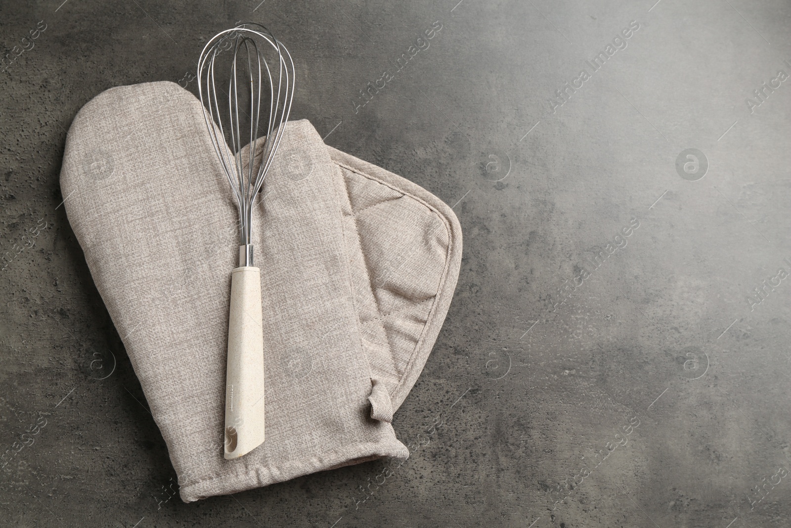 Photo of Grey oven glove, potholder and whisk on dark textured table, top view. Space for text