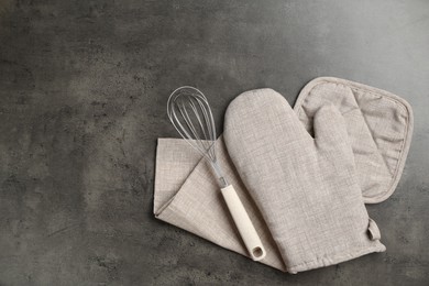 Photo of Grey oven glove, potholder, towel and whisk on dark textured table, flat lay. Space for text
