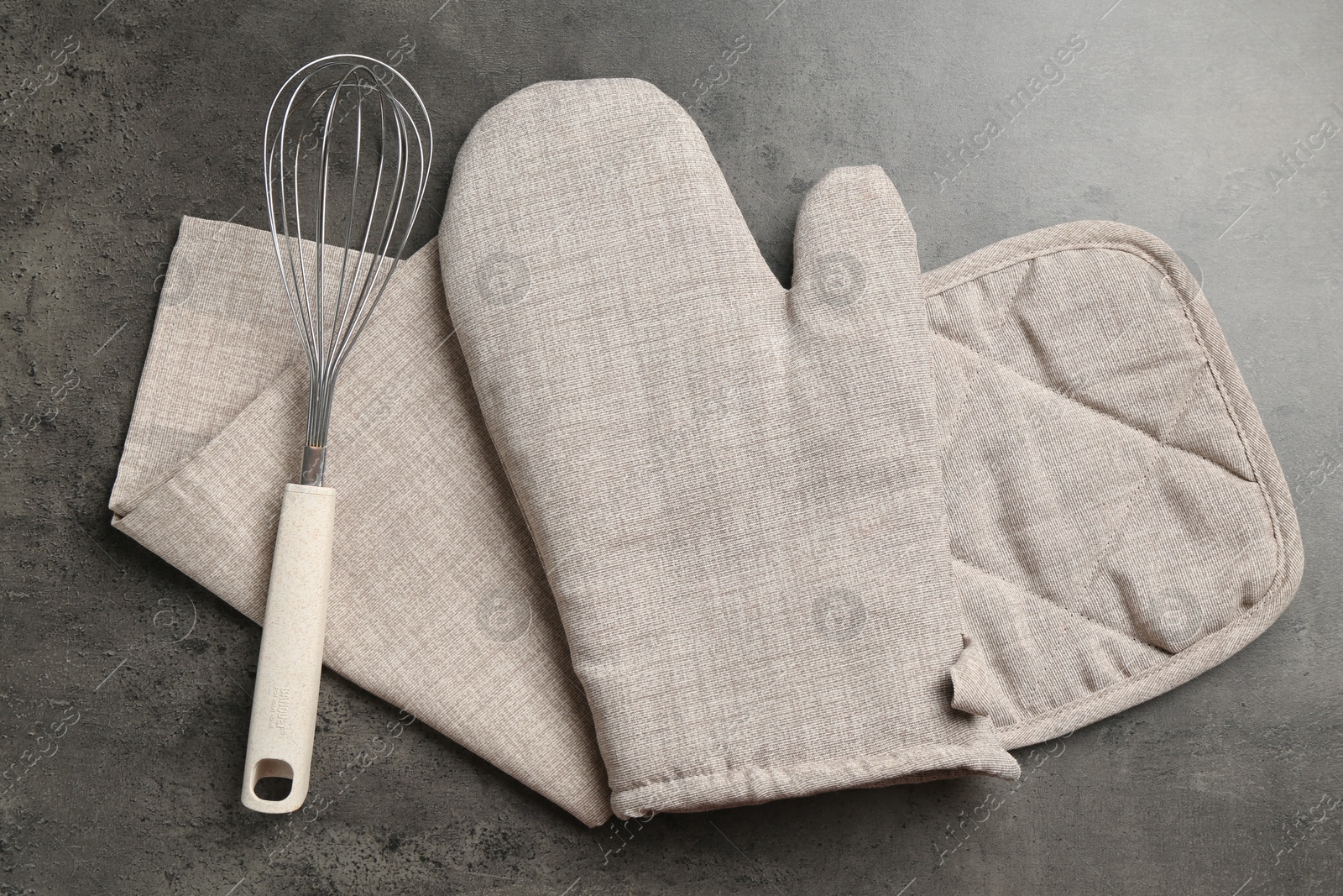 Photo of Grey oven glove, potholder, towel and whisk on dark textured table, flat lay