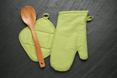 Photo of Green oven glove, wooden spoon and potholder on dark textured table, flat lay