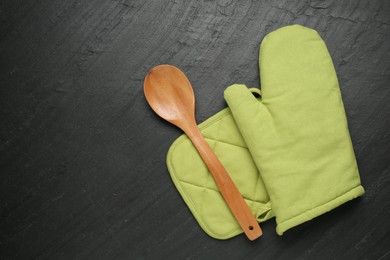 Photo of Green oven glove, wooden spoon and potholder on dark textured table, flat lay. Space for text