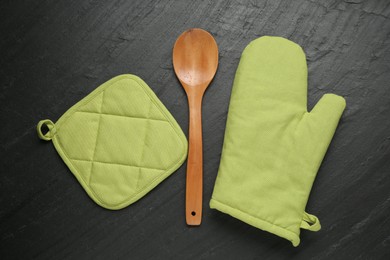 Photo of Green oven glove, wooden spoon and potholder on dark textured table, flat lay