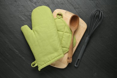 Photo of Green oven glove, wooden spoon, potholder, whisk and board on dark textured table, flat lay