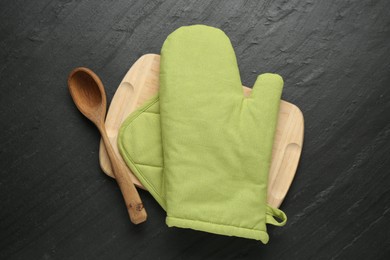 Photo of Green oven glove, wooden spoon, potholder and board on dark textured table, top view