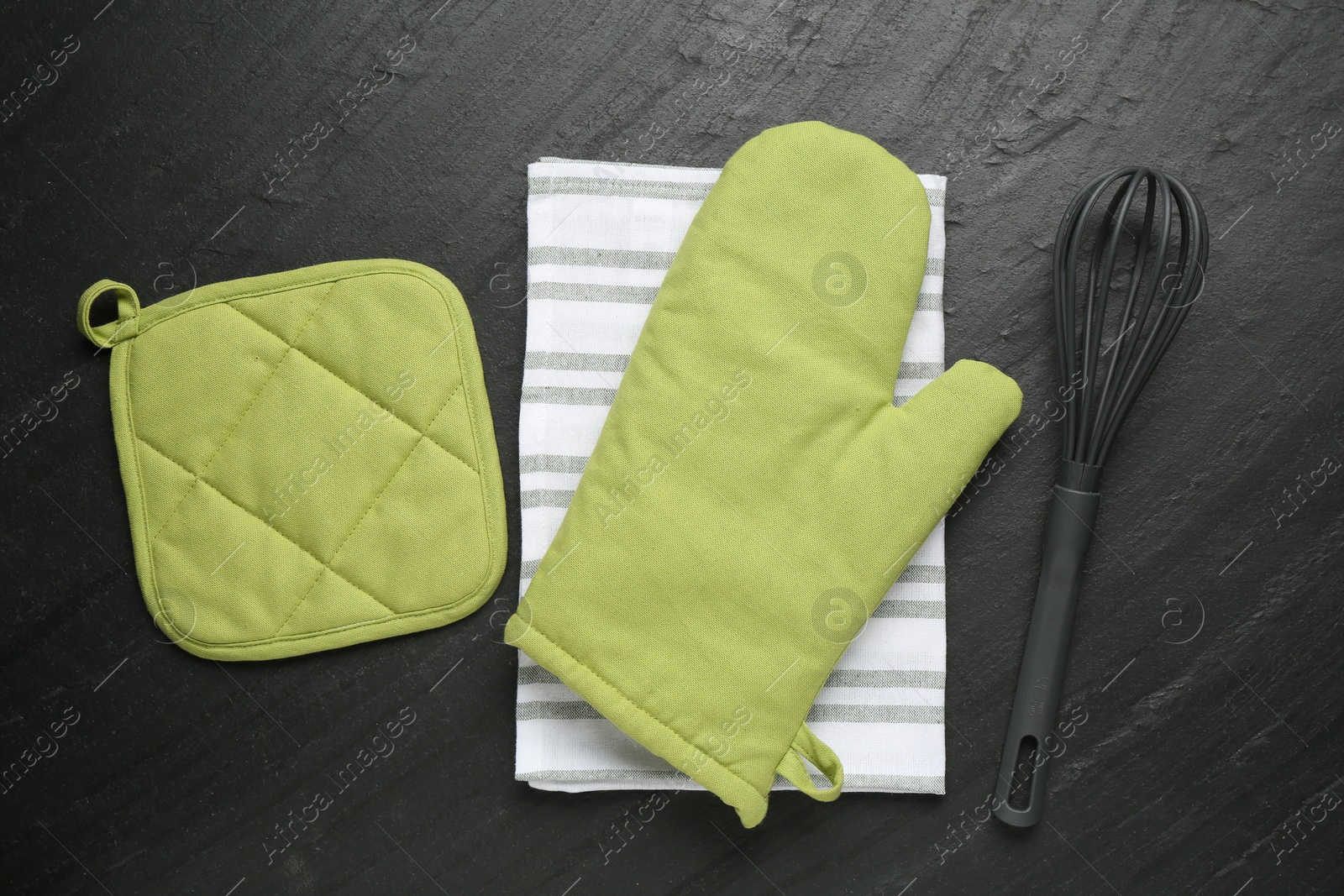 Photo of Green oven glove, towel, potholder and whisk on dark textured table, flat lay