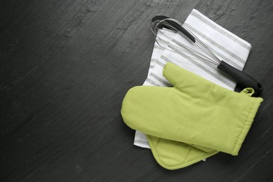 Photo of Green oven glove, towel, potholder and whisk on dark textured table, flat lay. Space for text
