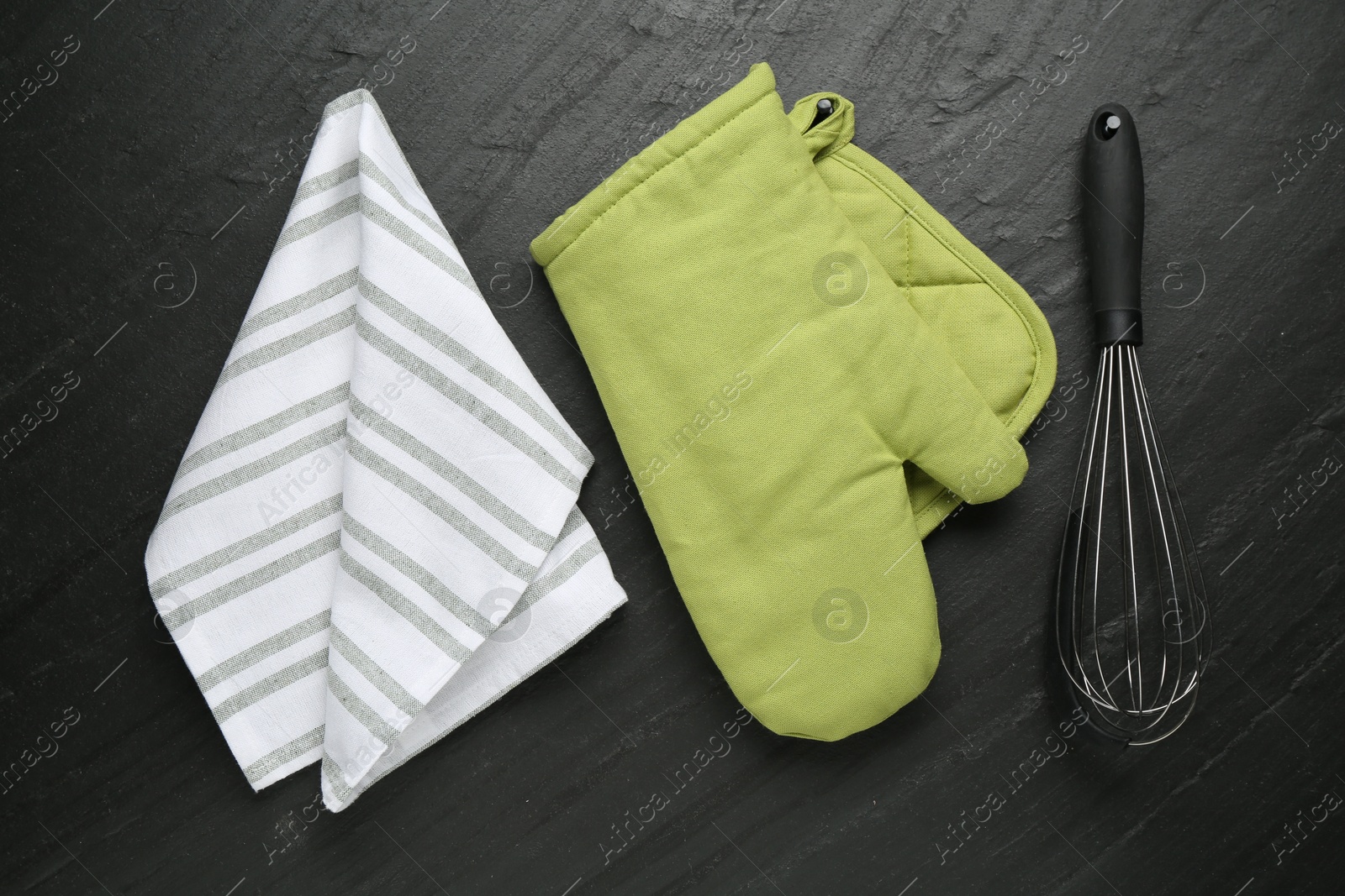 Photo of Green oven glove, towel, potholder and whisk on dark textured table, flat lay