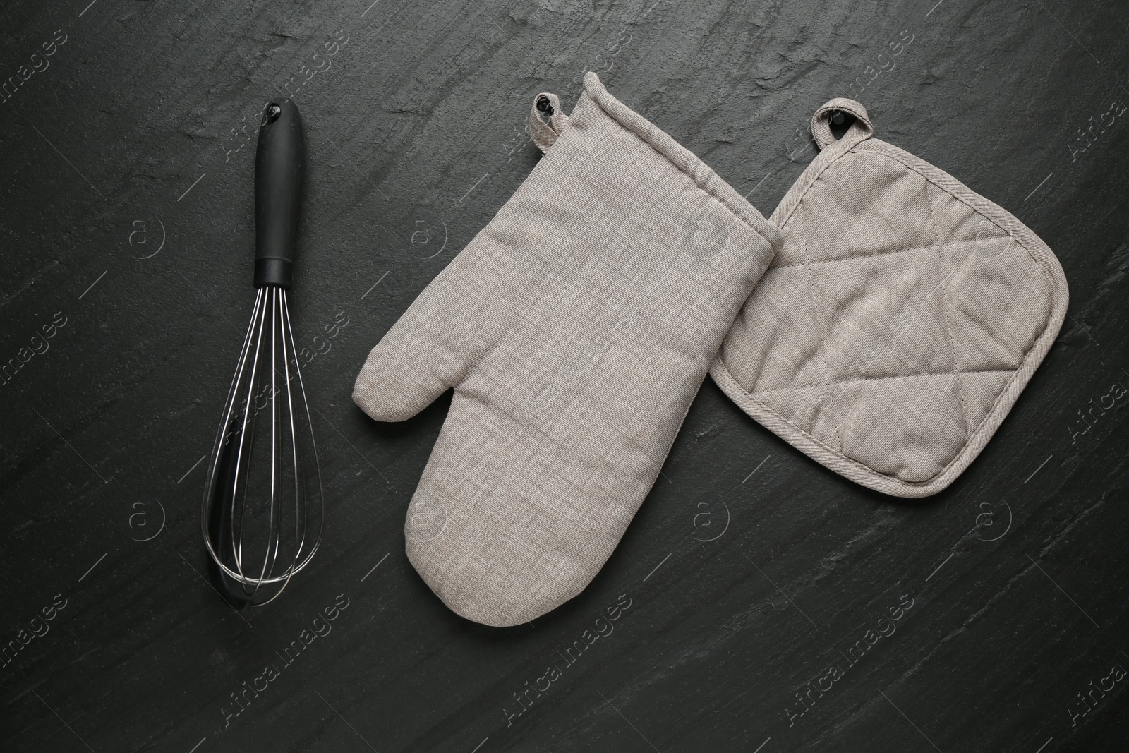 Photo of Grey oven glove, potholder and whisk on dark textured table, flat lay