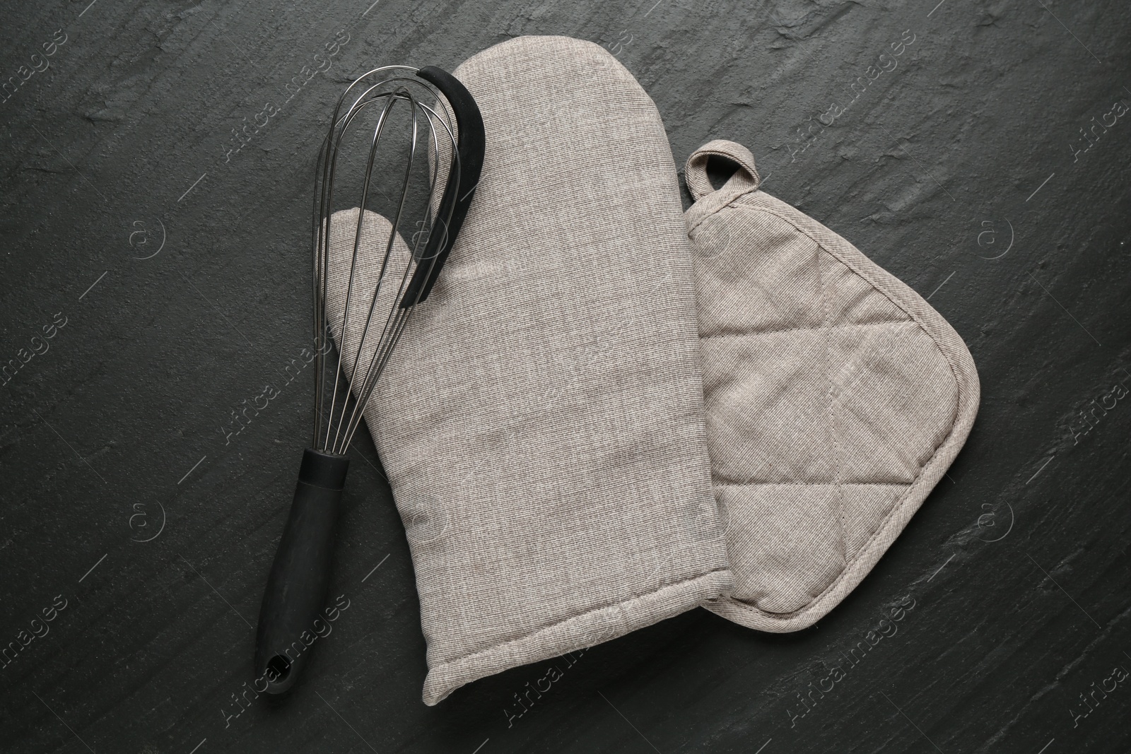 Photo of Grey oven glove, potholder and whisk on dark textured table, flat lay