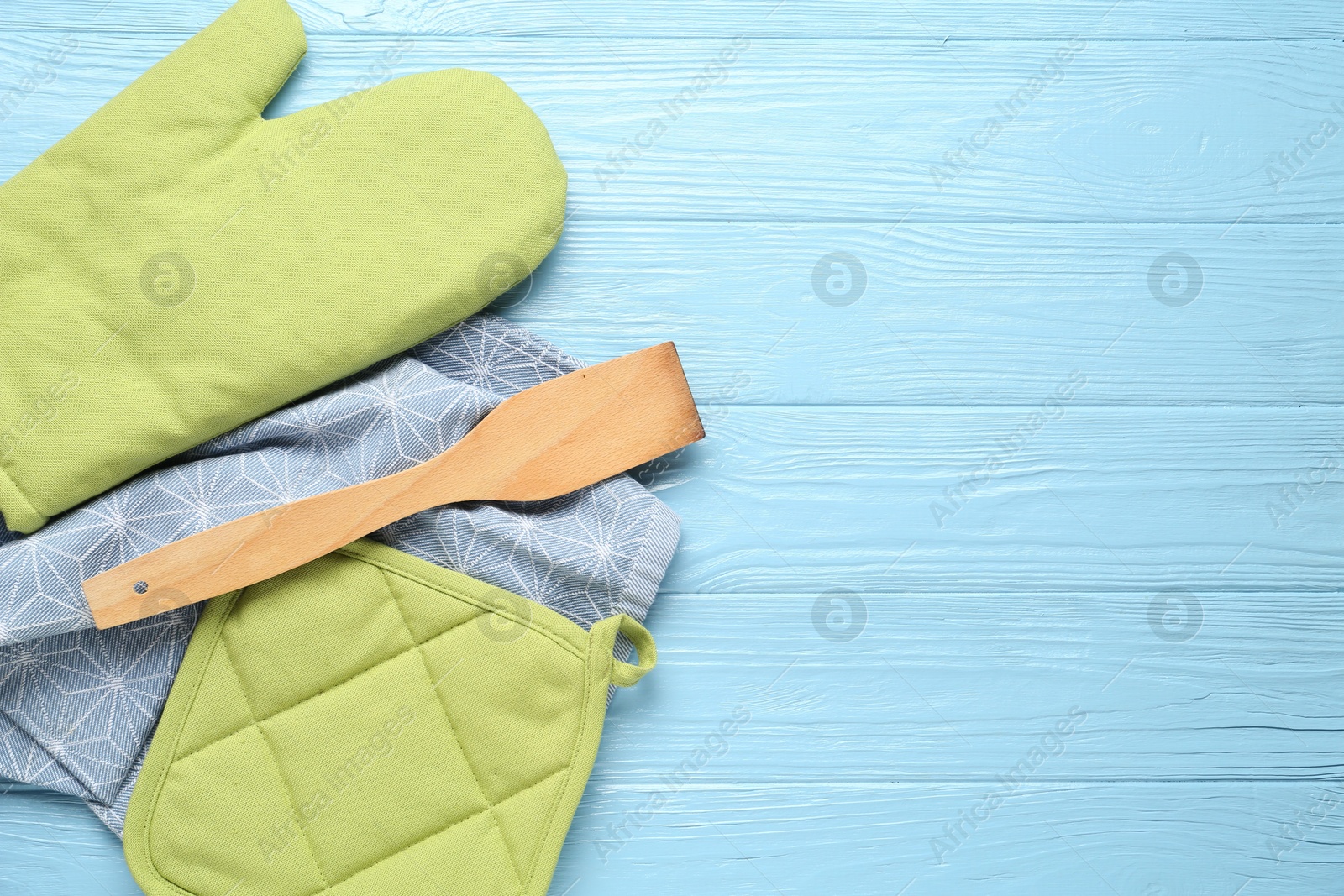 Photo of Green oven glove, potholder, towel and spatula on light blue wooden table, flat lay. Space for text