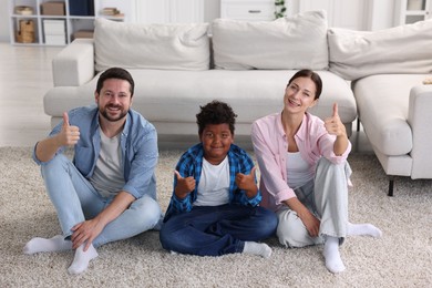 Photo of Adoption. Happy parents with their son showing thumbs up on floor at home