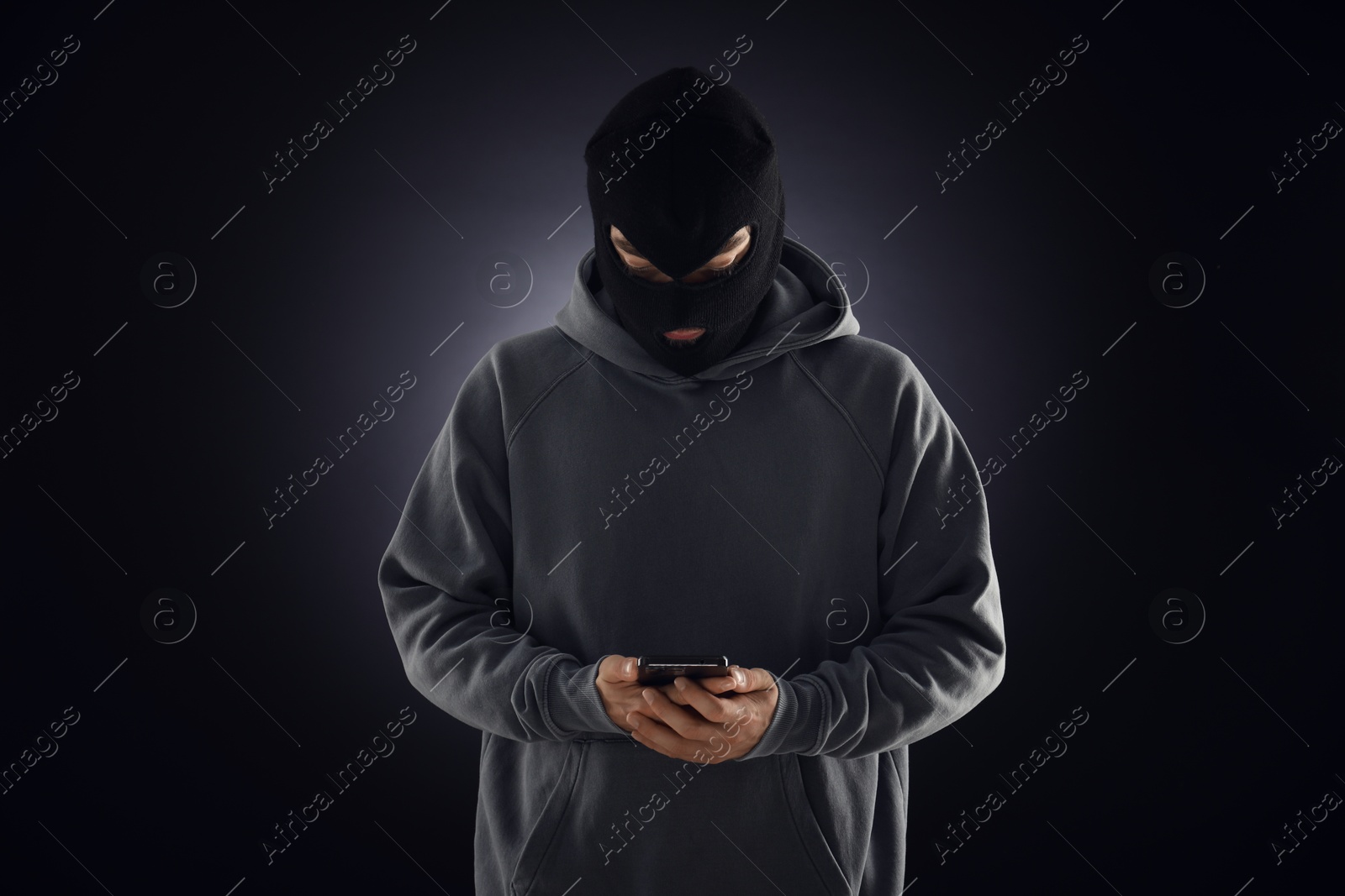 Criminal in mask with phone against dark background Photo of Criminal in mask with phone against dark background