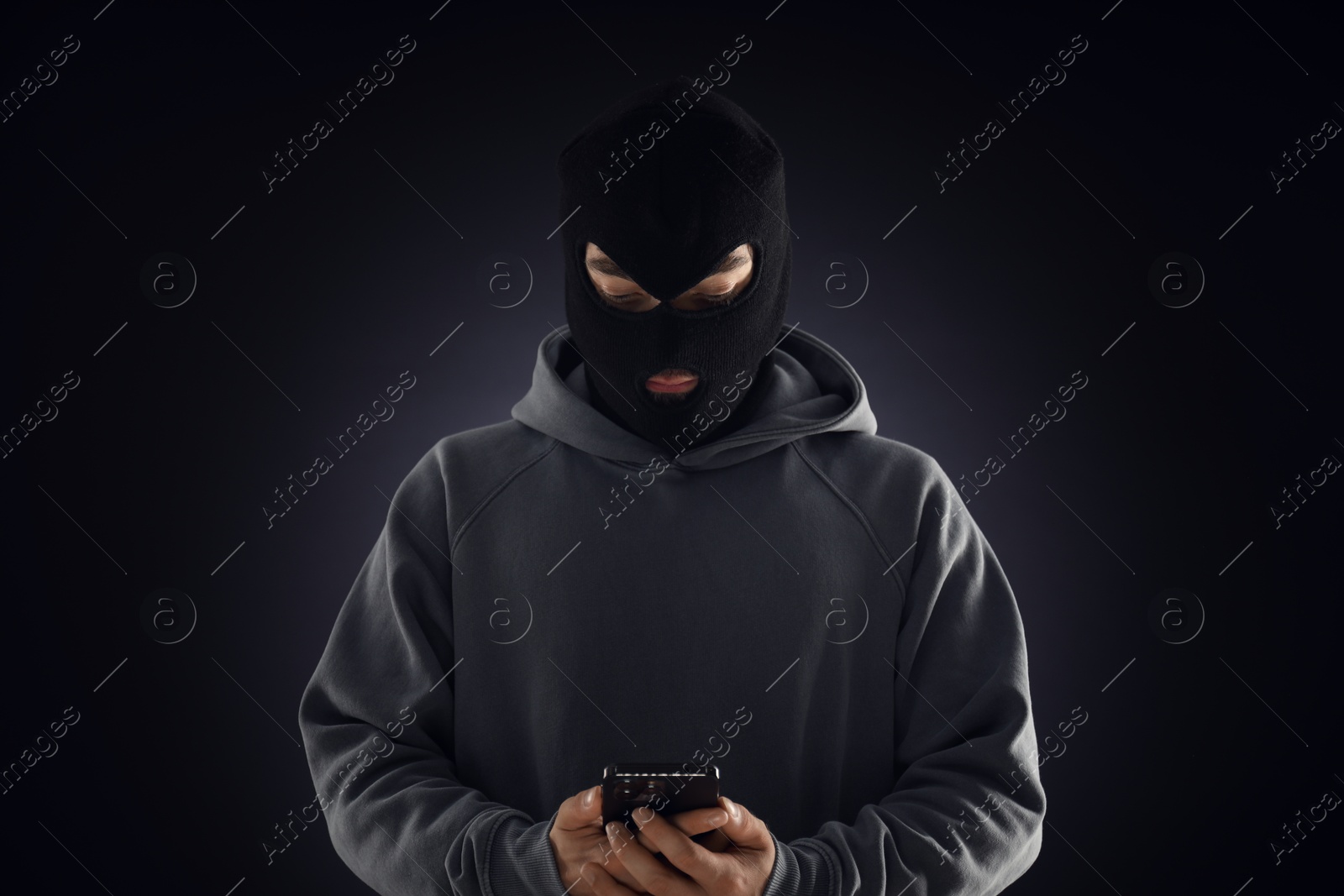 Criminal in mask with phone against dark background Photo of Criminal in mask with phone against dark background