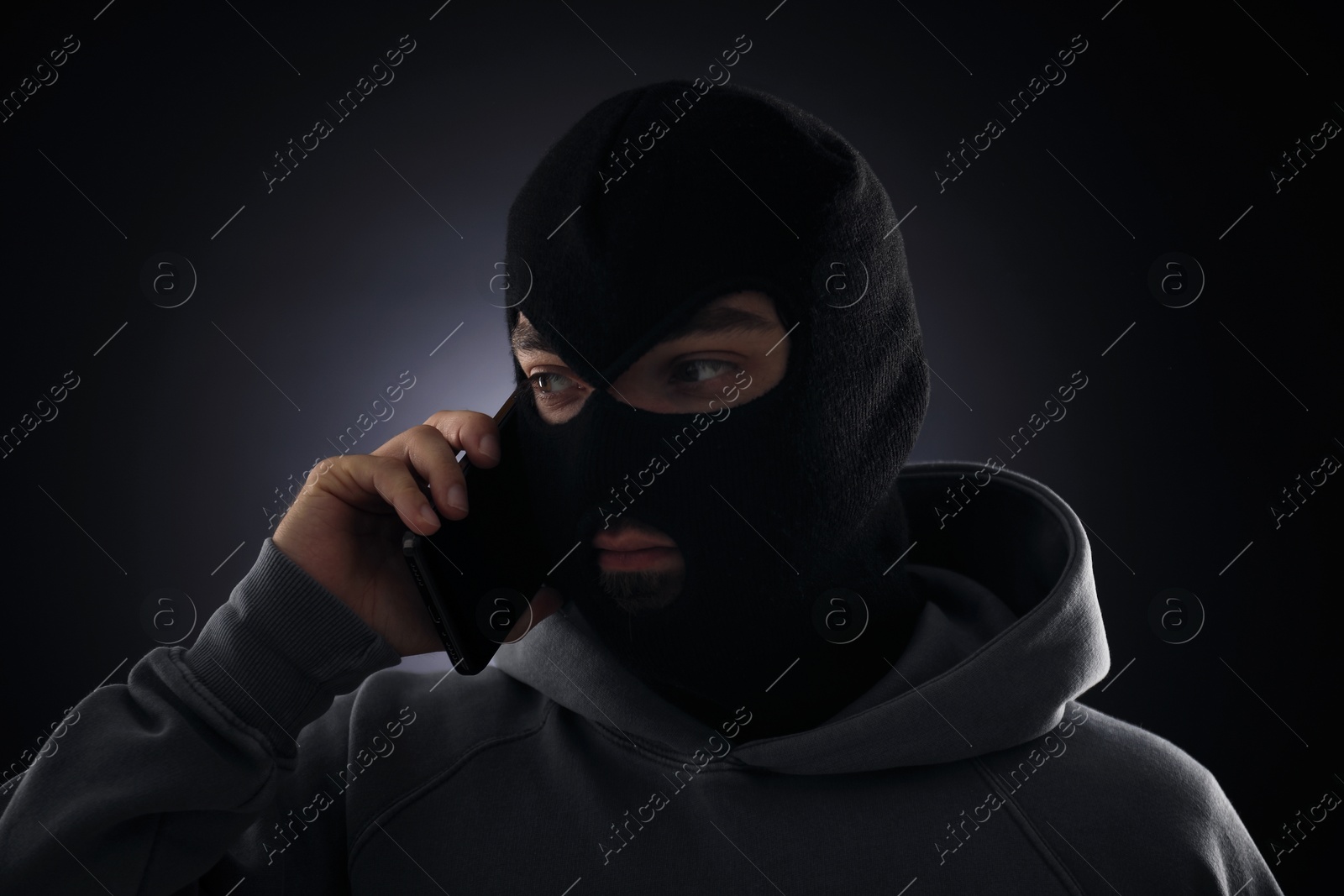 Criminal in mask talking on phone against dark background Photo of Criminal in mask talking on phone against dark background