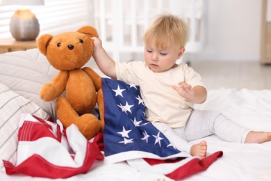 Photo of Cute little child with flag of USA and toy bear on bed at home