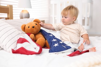 Photo of Cute little child with flag of USA and toy bear on bed at home