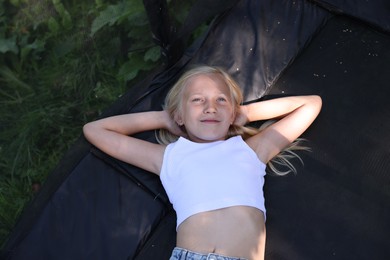 Photo of Cute little girl lying on black trampoline outdoors, top view