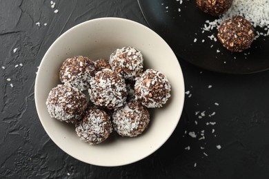 Photo of Tasty chocolate candies with nuts and coconut flakes in bowl on black table, flat lay