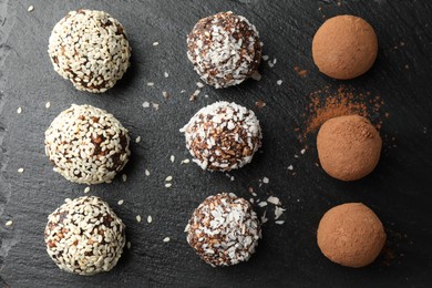 Photo of Tasty chocolate candies with cocoa powder, nuts, coconut flakes and sesame seeds on black table, flat lay